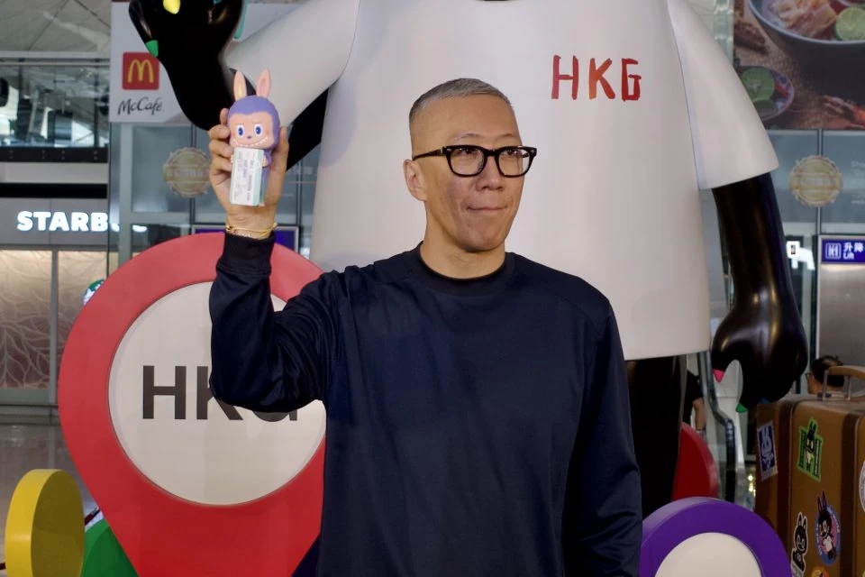 Un uomo mostra un piccolo Labubu davanti a un'installazione Pop Mart in aeroporto a Hong Kong