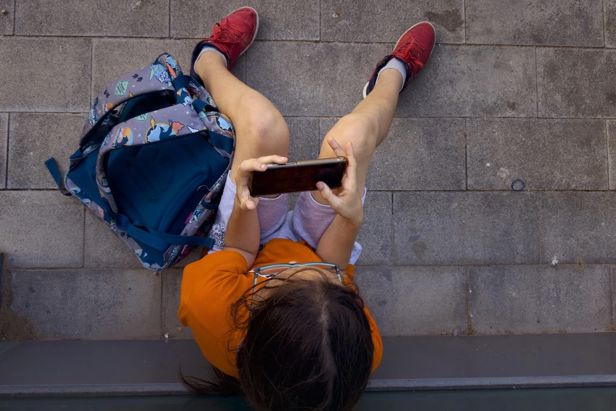 Un bambino seduto a terra con lo zaino, assorto nello smartphone