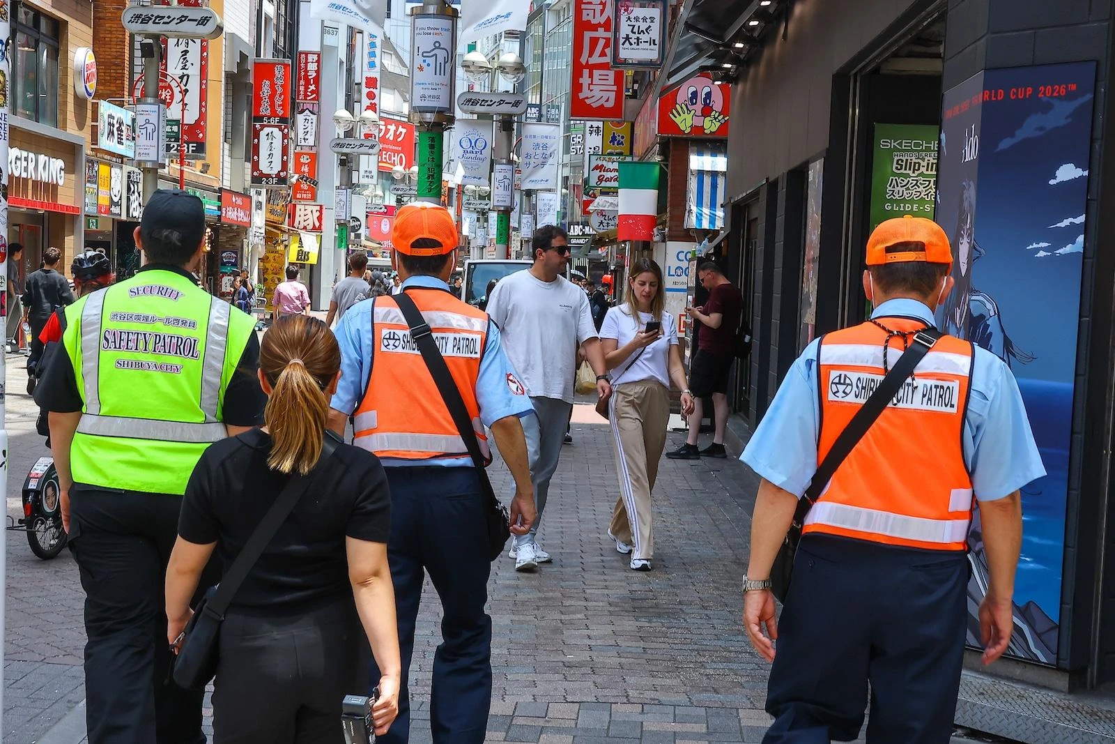 Addetti della Shibuya City Patrol in gilet fluo e arancione pattugliano una via dello shopping affollata